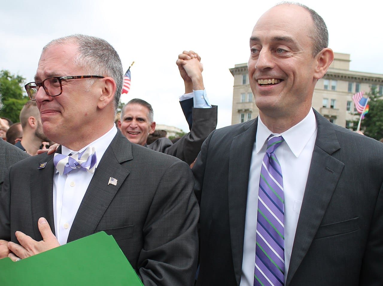 A tearful Jim Obergefell in a suit and purple bow tie, weeps joyfully as he stand beside his attorney and in front of a jubilant crowd.