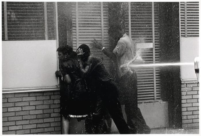 Three Black protesters up against a brick wall and glass windows are hit by a high pressure stream of water and clearly in great discomfort.