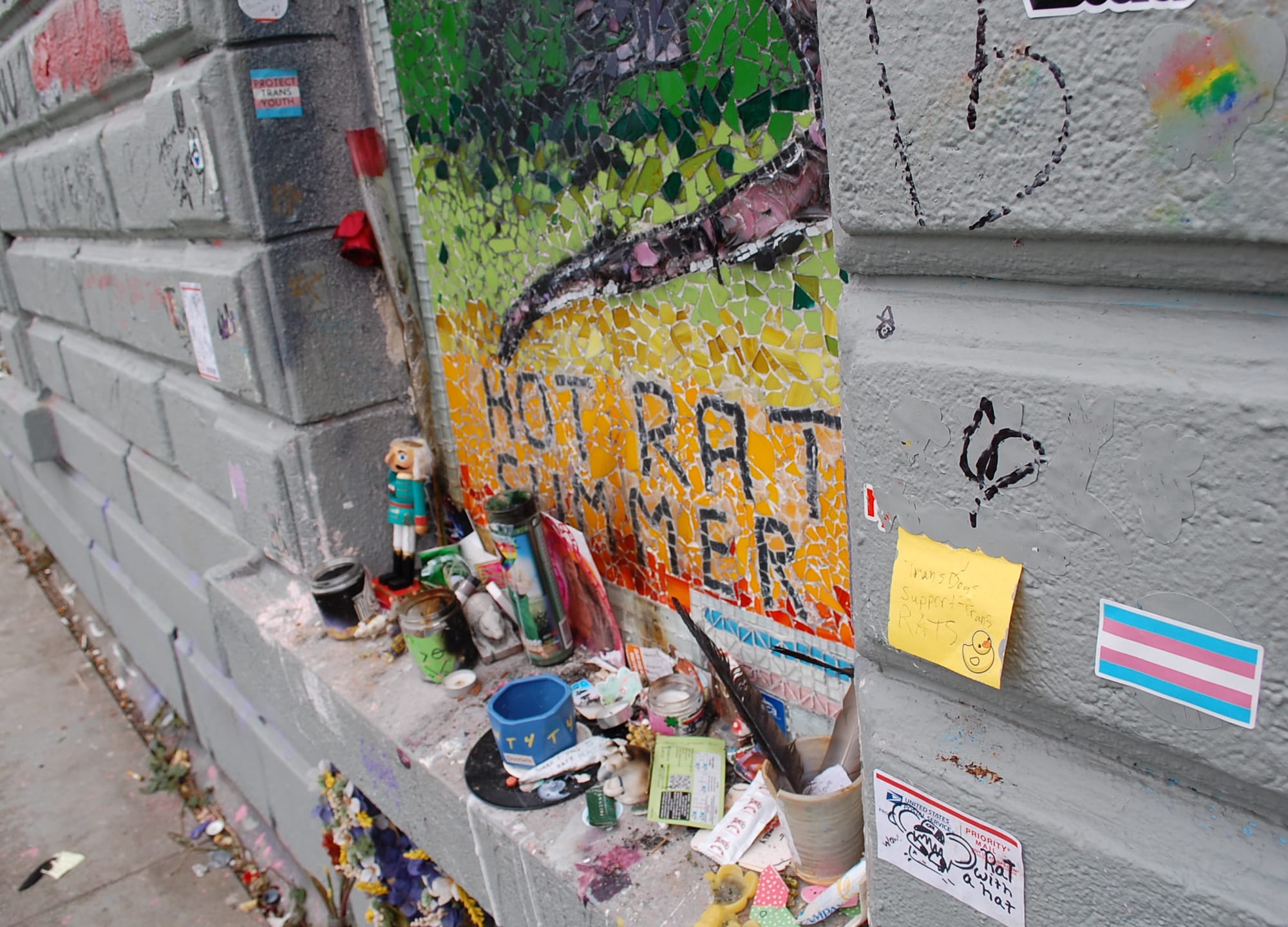 A mosaic that reads "Hot rat Summer" is adorned with offerings, candles, feathers, and trans flags.