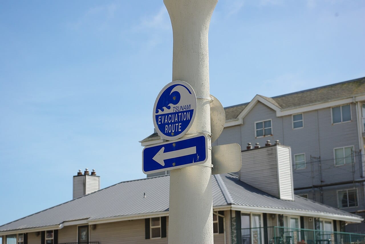A blue and white tsunami evacuation route sign points to the left in front of a three story apartment building.