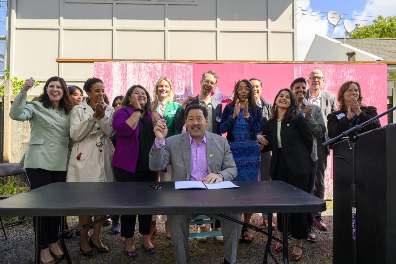 Bruce Harrell lifts a pen and smiles with a crowd of people behind him.