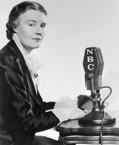 A woman reporter in a jacket and sweater sits in front of an old-time microphone with the logo of NBC on the side