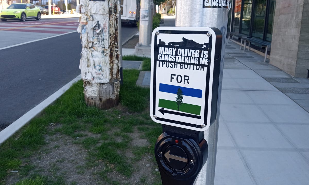A "beg button" requesting a walk sign at a Seattle crosswalk is decorated with stickers, including a Cascadia Flag and "Mary Oliver is Gangstalking Me"
