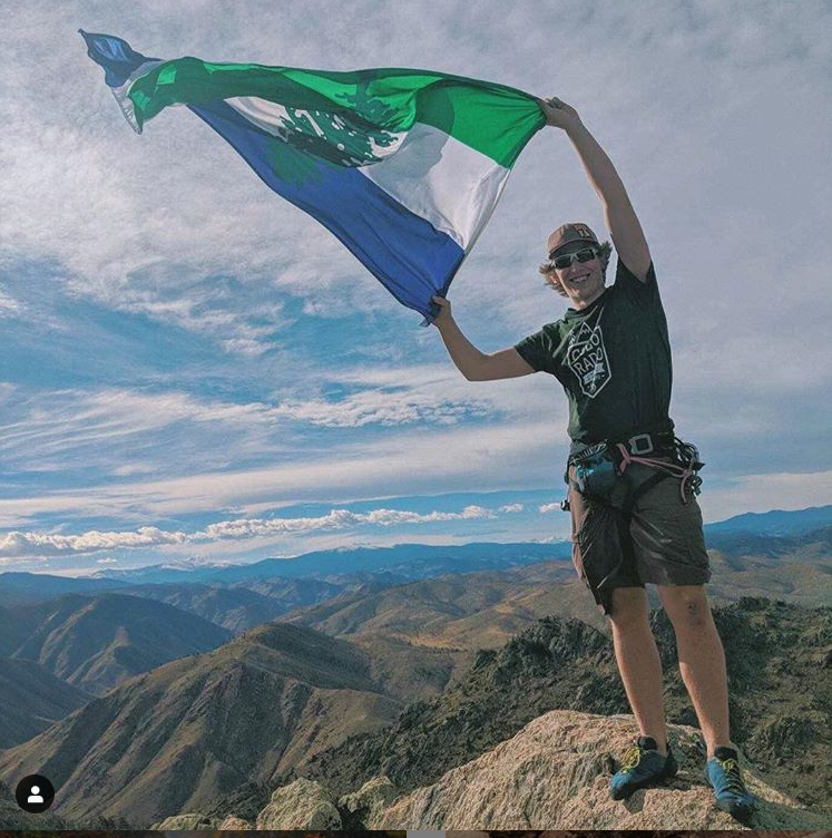 A hiker holds a Cascadia Doug flag in the wind at the top of a peak in the Cacade range.