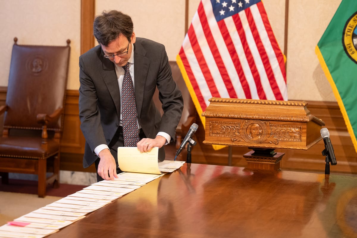 Washington governor Bob Ferguson looks at a stack of bills lined up to sign on a desk, with the flags of the United States and Washington in the background.