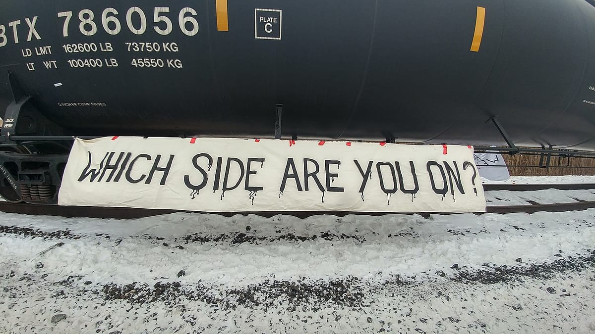 A sign reading "WHICH SIDE ARE YOU ON" is displayed below a tanker car on an train carrying liquified natural gas.