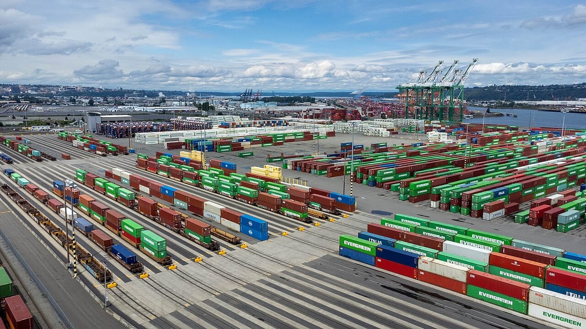 A view of the Port of Tacoma on a cloudy day, with red, blue, and green shipping containers lined up waiting to be put on trains in the foreground, cranes in the background.