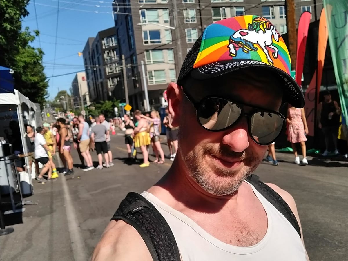 A man in a tank top, mirrored sunglasses and a rainbow unicorn trucker hat stands on a street with others celebrating Pride on a Seattle street.