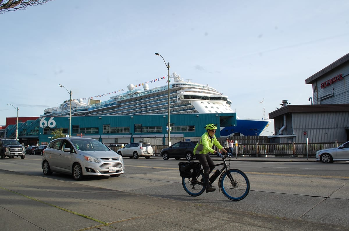 On the Seattle waterfront, a large cruise ship is docked at pier 66 as a car and a cyclist pass in front of it.