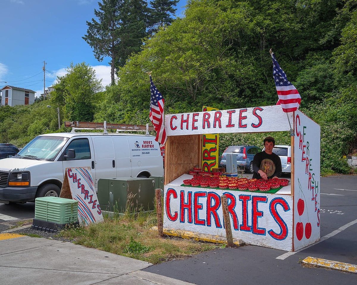 A young man sells cherries at a small roadside stand decorated with American flags.
