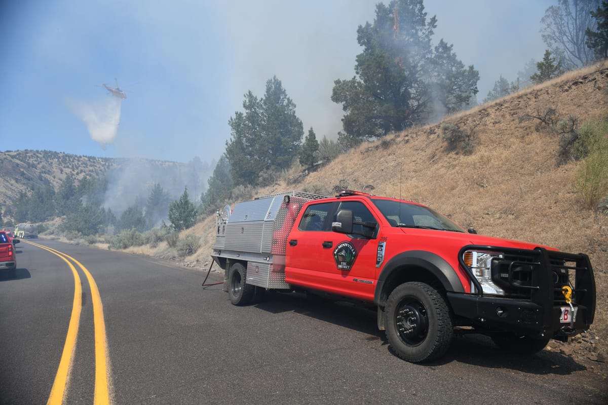 A forest fighting pickup truck is seen on the side of a highway as a helicopter drops water on a fire among scattered pine trees.