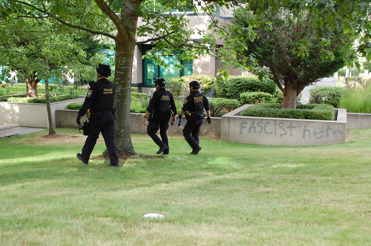 Three DHS officers in black tactical gear and helmets walk across a lawn next to a wall spray-painted with the words "FASCIST HERE."