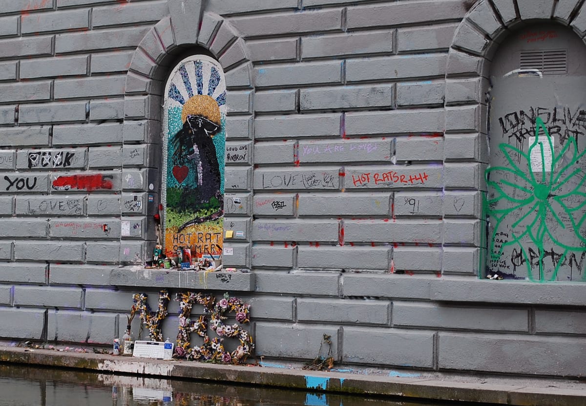 A mosaic of a rat, with rising sun and adorned with trans flags, candles, and a sculpture that reads YES.