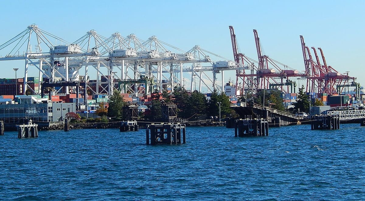 A dozen white and red cranes stand above ships and shipping containers with blue waters of Eliott Bay in the foreground.