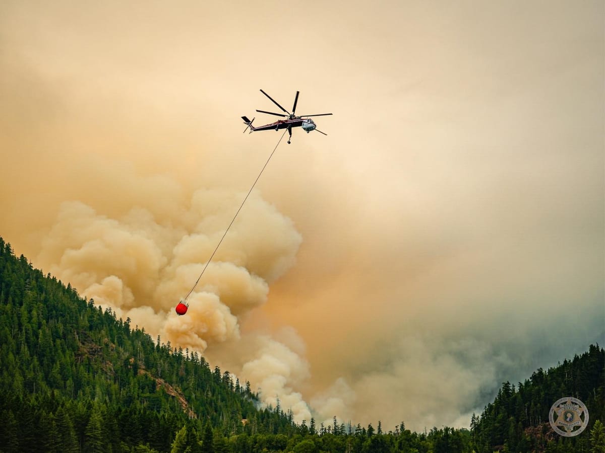 A helicopter drops water on a forest fire as smoke billows in the background.