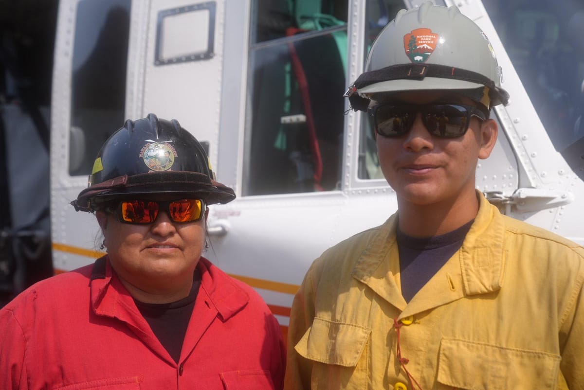 Two firefighters wearing helmets standing in front of a helicopter, including one with the logo of the National Park Service.