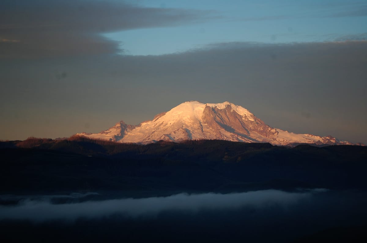 The glaciers of Tahoma, also known as Mount Rainier, glow in early morning light with foggy mountains in the foreground.