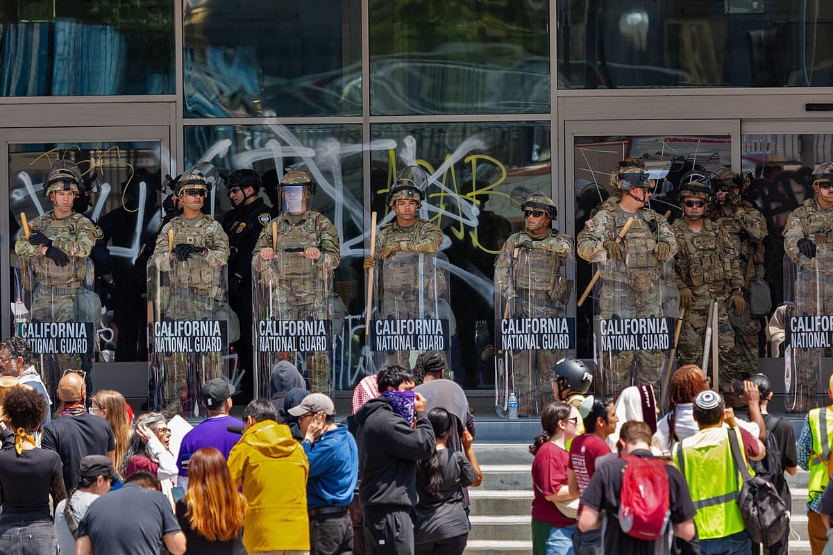 Protesters stand below the steps of a building lined with National Guar troops holding batons and clear plastic riot shields.