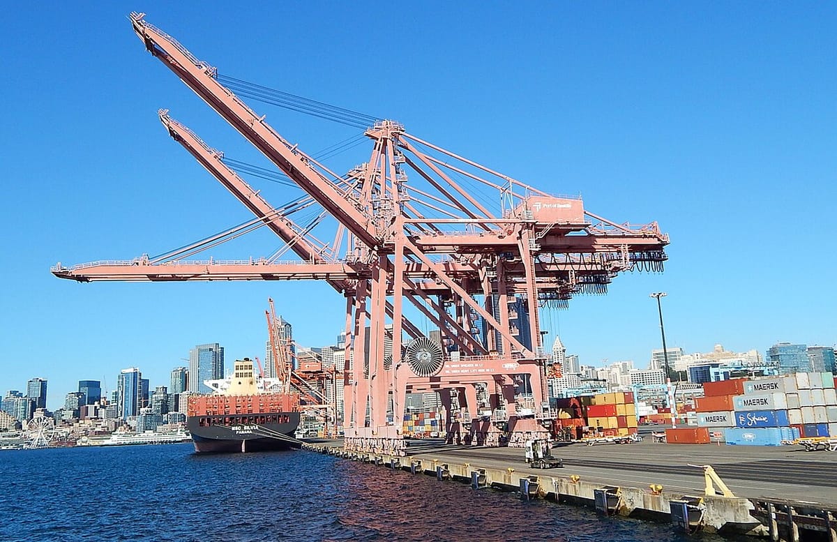 Large cranes load and unload containers above a cargo ship, with the Seattle skyine in the background.