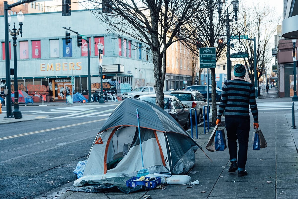 A person in winter clothes and shopping bags walks base a tent used by someone who is homeless.
