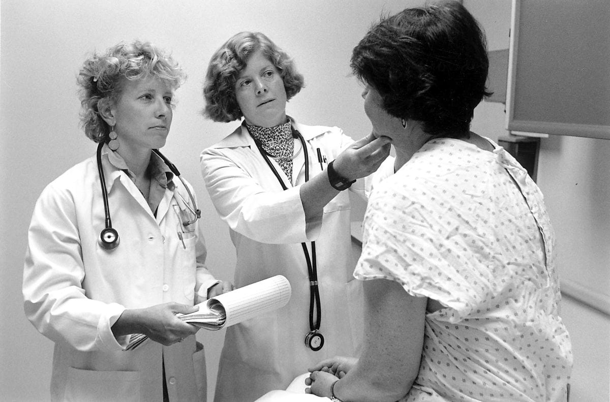 Two women doctors with stethescopes and white uniforms examine a female patient in a hospital gown