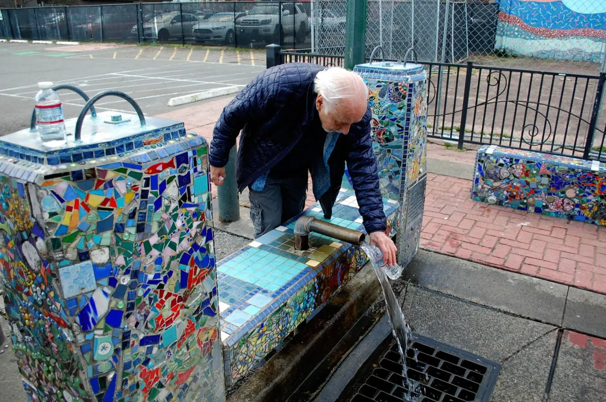 An elderly man collects water in a bottle from a fountain decorated with colorful mosaics.