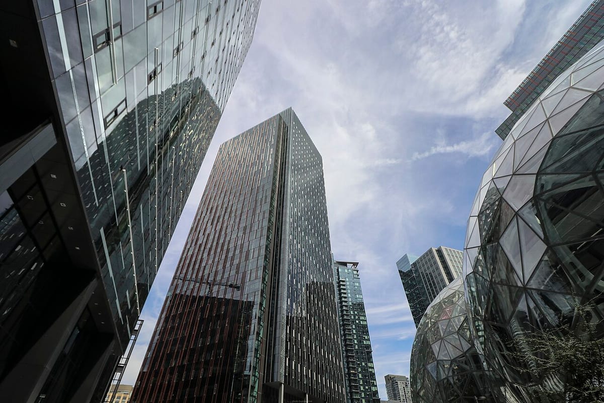 Several tall skyscrapers and a cloud filled blue sky next to the geodesic spheres at Amazon's Seattle headquarters.