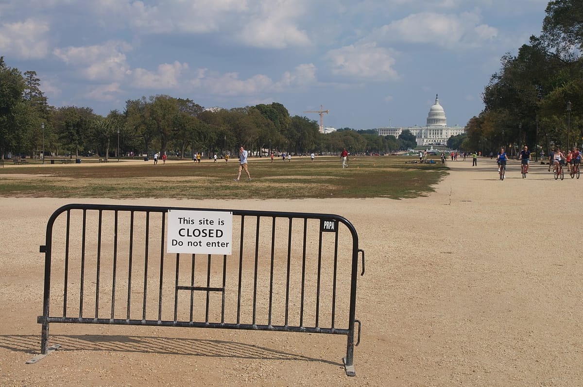 A single barrier stands in the National Mall with the US Capitol building in the distance. On the barier a sign reads This site is closed, do not enter while multiple people walk there anyway