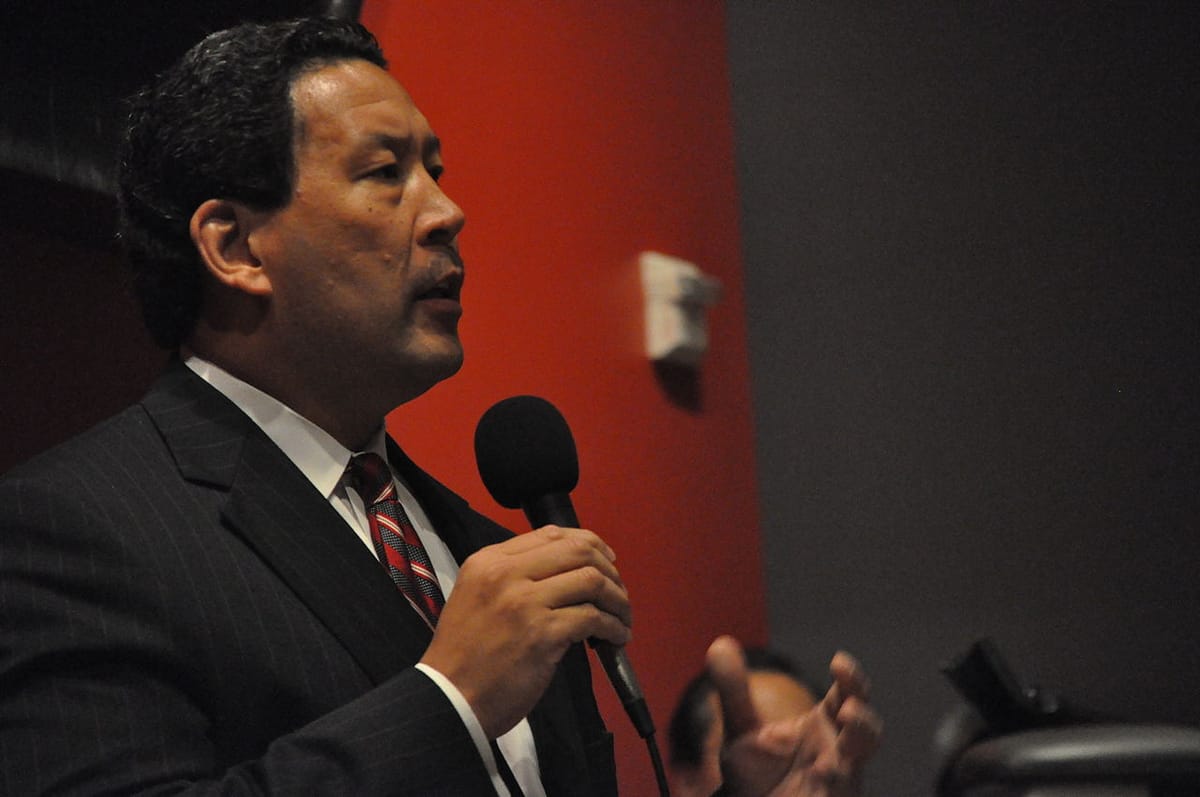 Bruce Harrell speaks into a microphone with a dark red wall behind him.