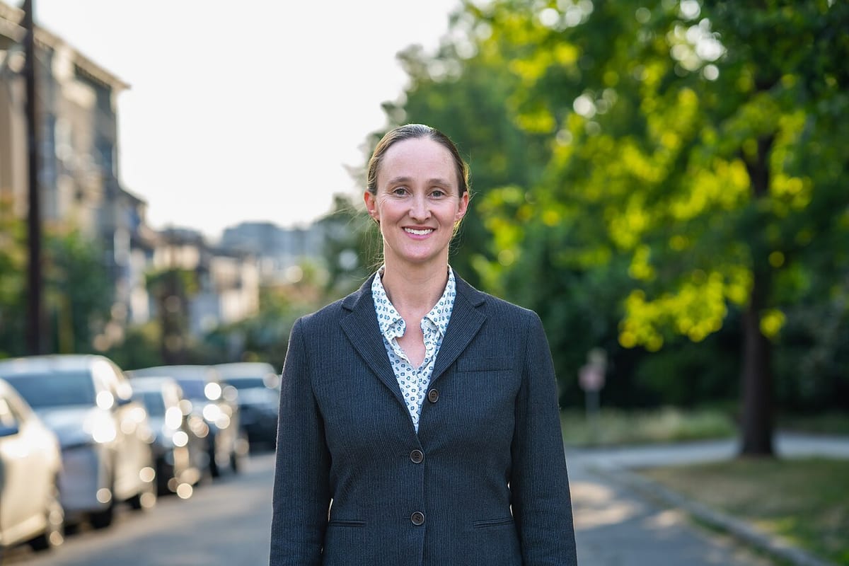 Seattle mayoral candidate Katie Wilson stands on a wooded Seattle street wearing a casual business suit.
