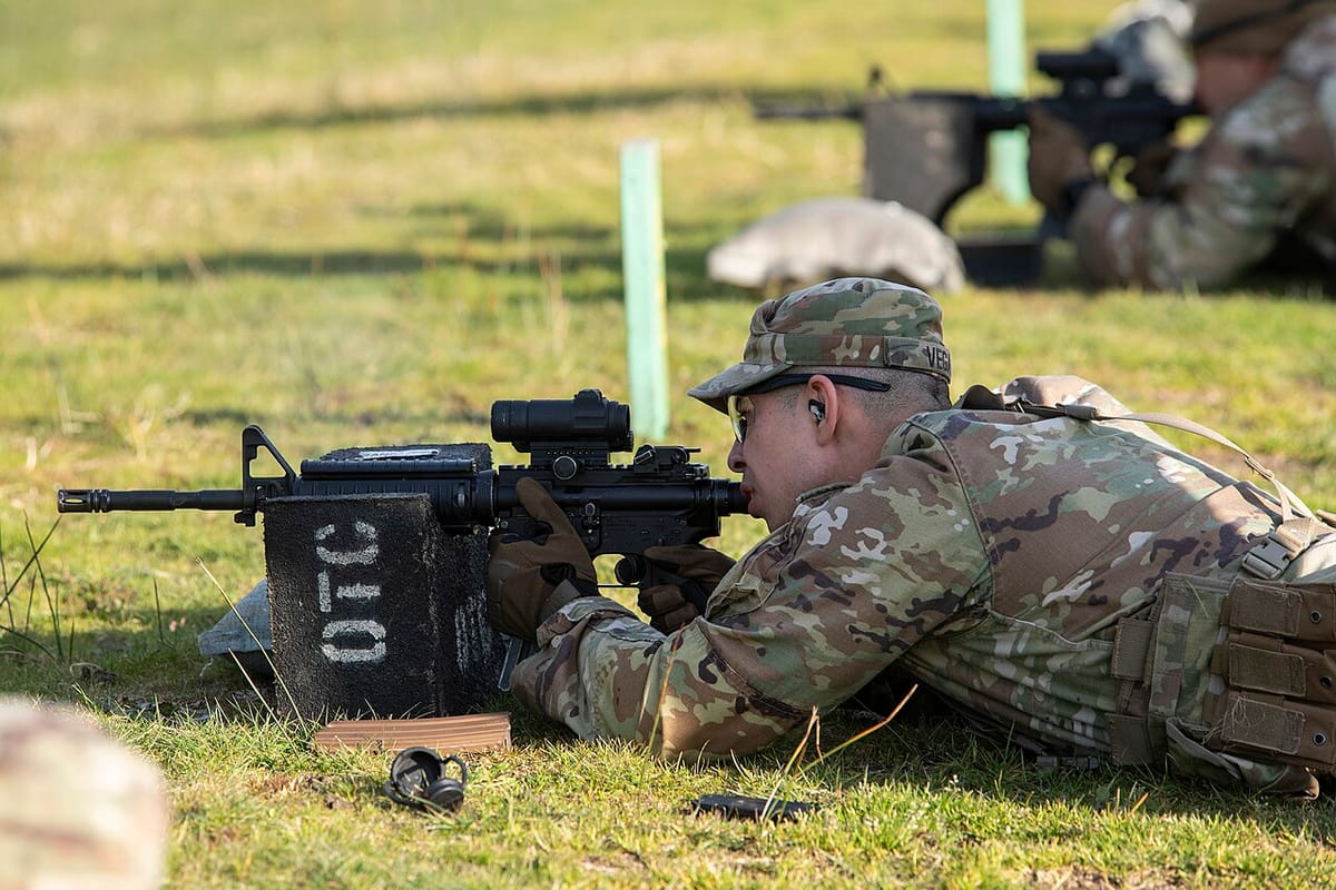 A soldier in camoflauge lying on the ground aims an assault rifle.
