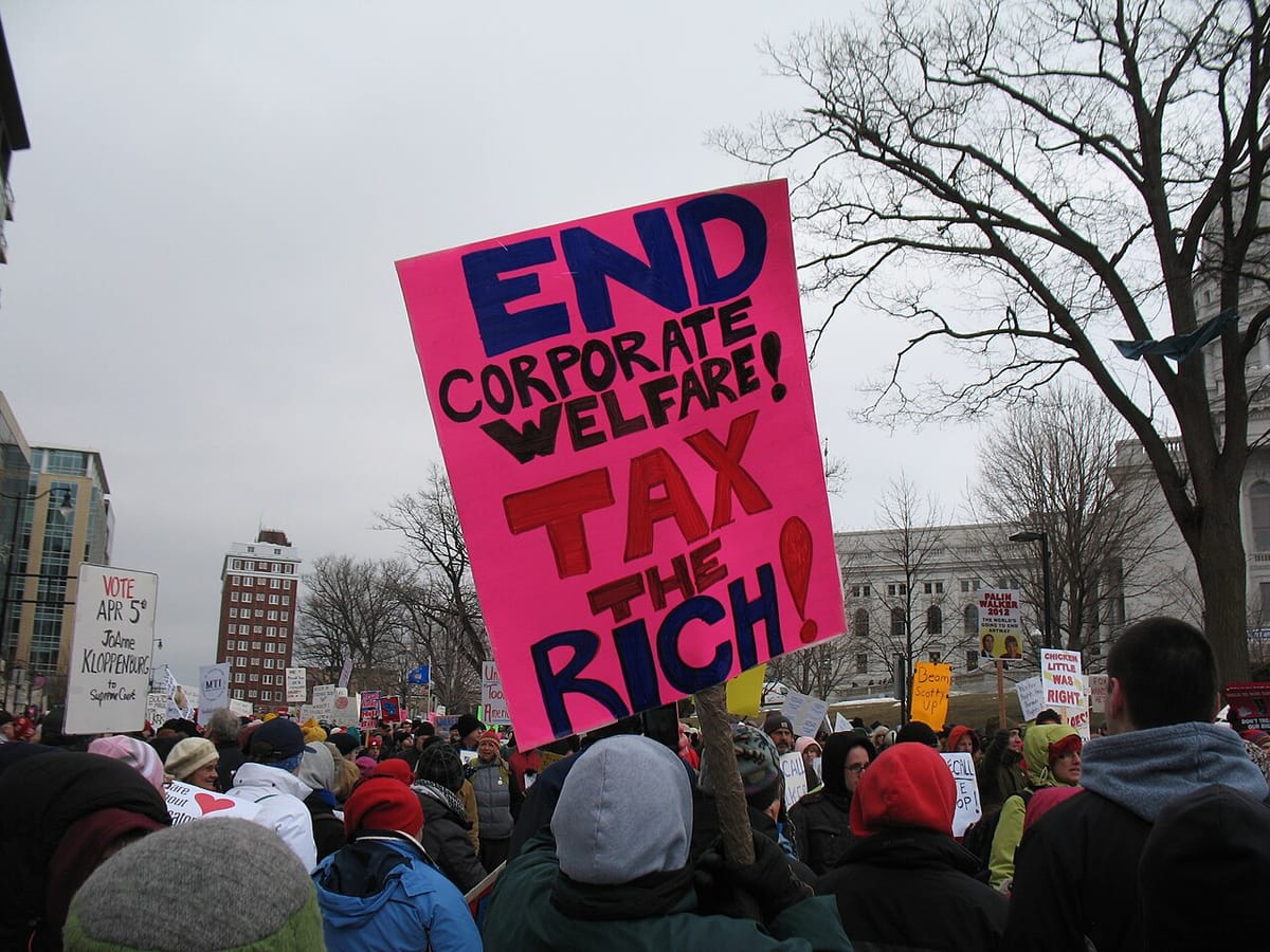 A crowd of people protesting, with one holding a sign that reads: End Corporate Welfare! Tax the Rich!