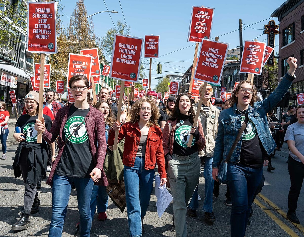 Starbucks workers in union t-shirts march down a street carrying signs that read Demand Starbucks Stop Cutting Employee Hours