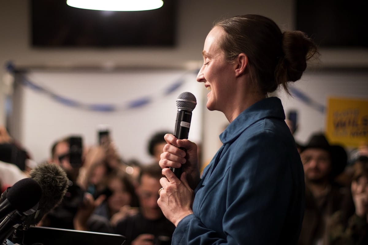 Katie Wilson speaks to a crowd, smiling, her eyes closed and gripping a microphone with two hands.