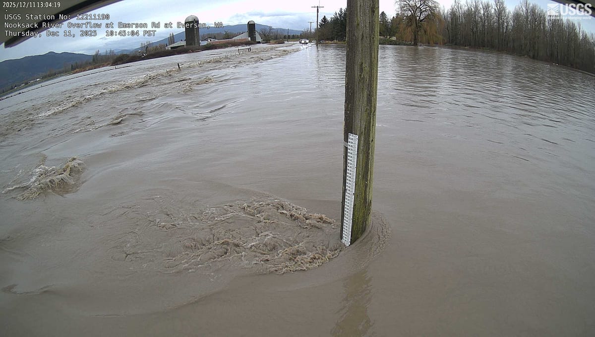 A remote camera view showing brown floodwaters, a rain gauge, and farm buildings in the background 