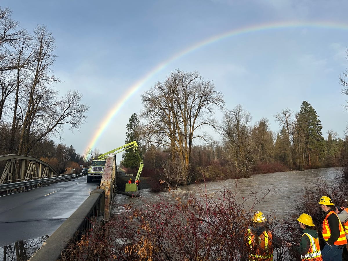 A rainbow shines over a highway bridge, with workers using a crane to pull debris from a flooded river