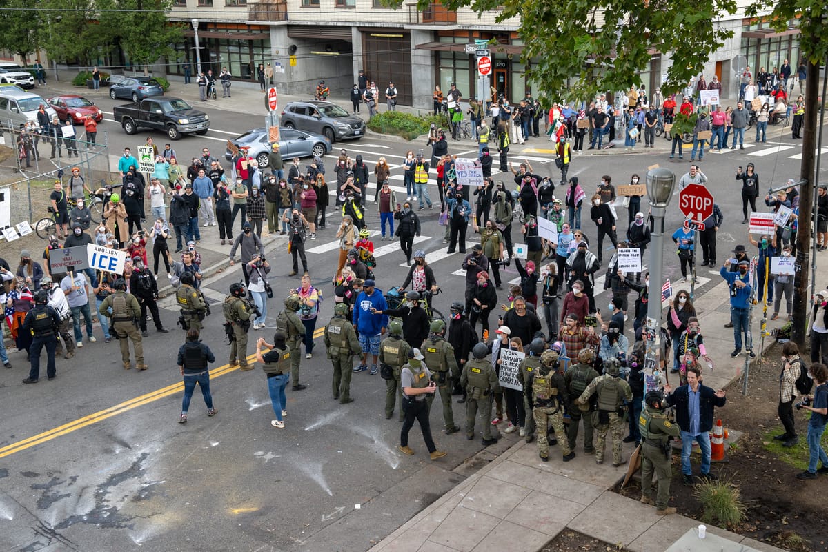 A crowd of protesters, seen from above, face down armed goon sin military gear. 
