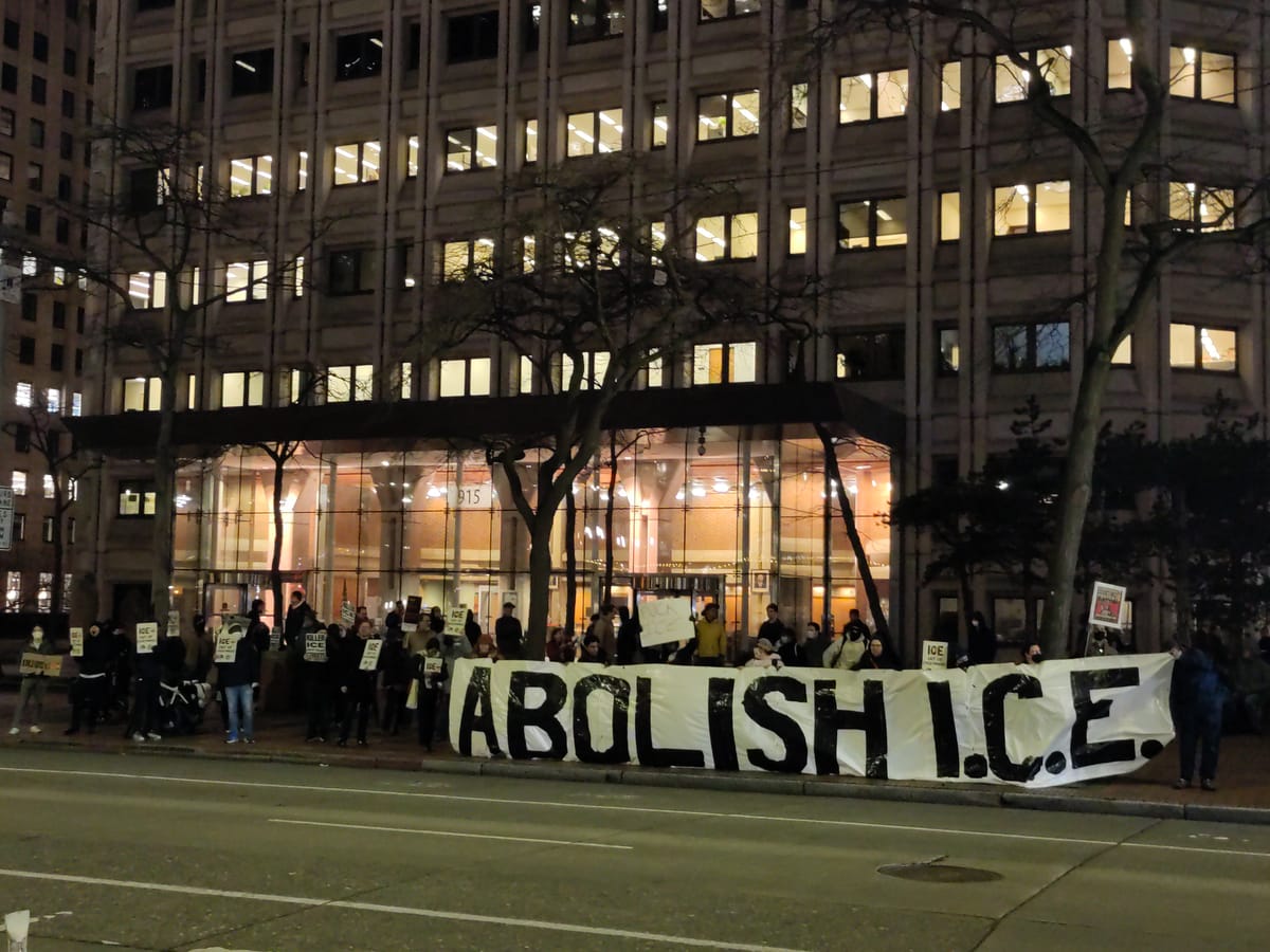 Protesters stand in front of an office building at night holding signs and a large banner that reads: Abolish ICE