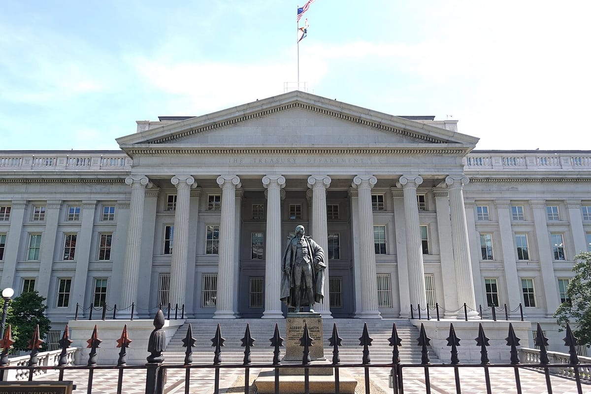 The facade of the US Treasury building with Greek columns, a statue of a revolutionary era figure and an iron gate.