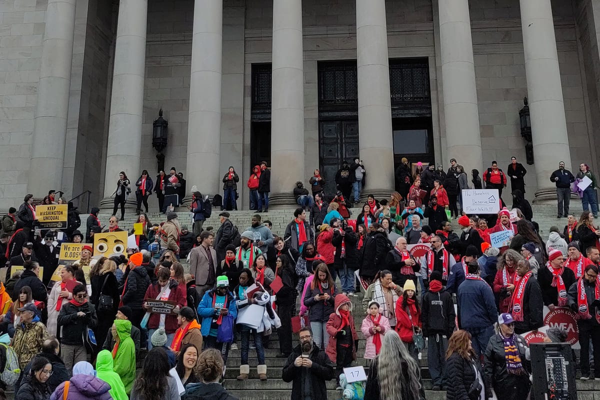 A crowd of people wearing red scarves and holding signs supporting raising taxes on the wealth stand on the steps of the legislative build