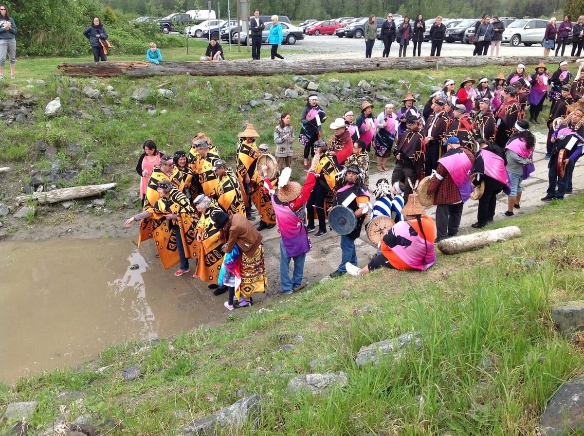 Indigenous people in colorful robes and tunics and some wearing woven cedar hats stand on a shoreline