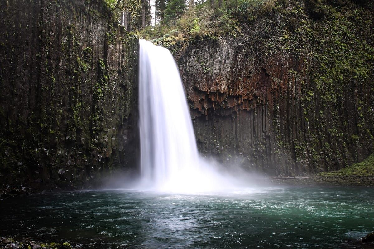 An elegant waterfall cascades into a deep green pool surrounded by walls of striated dark basalt.