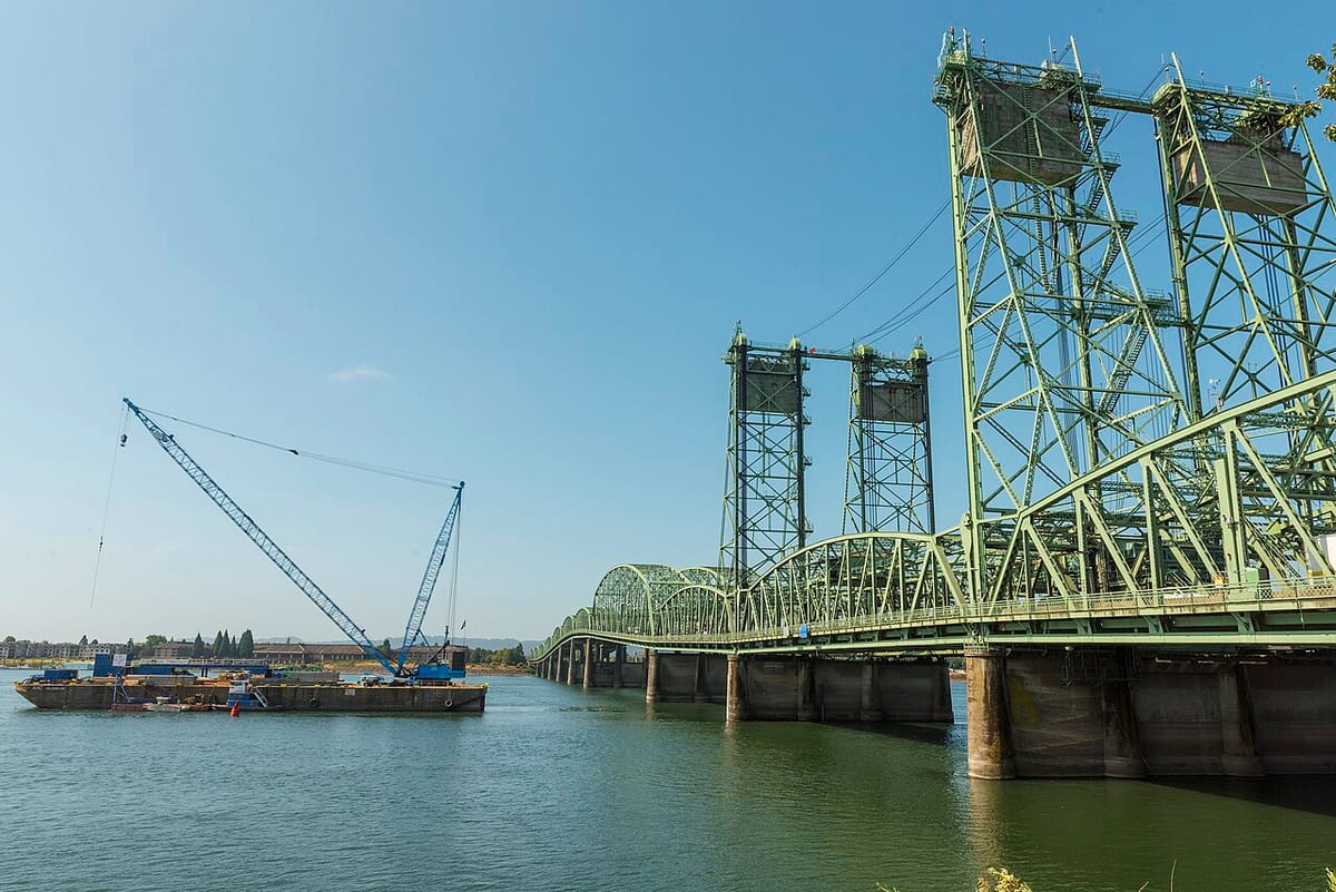 A long, cantelievered freeway bridge over a river, with a barge next to it.