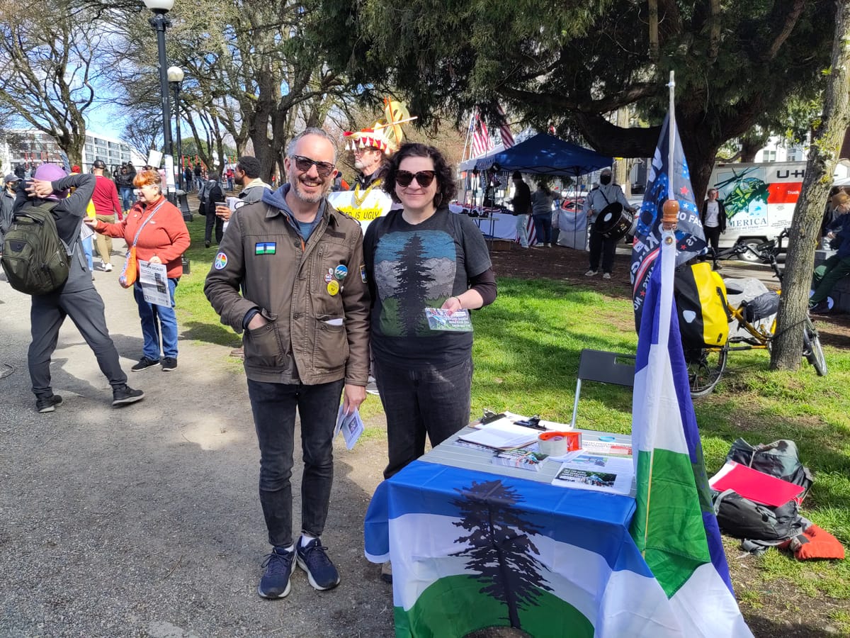 Two people wearing Cascadia themed clothing stand next to a table with flyers and a Cascadia flag