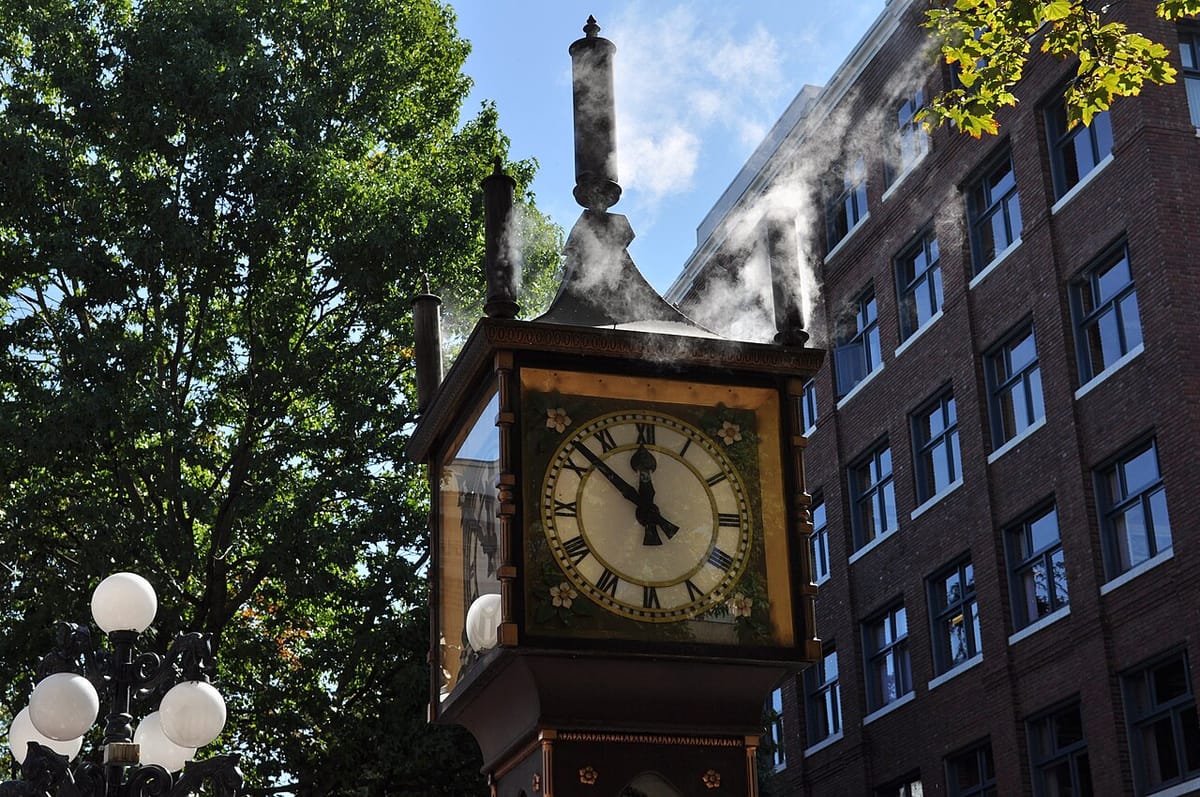An old fashioned outdoor clock emits steam from an exhaust whistle.