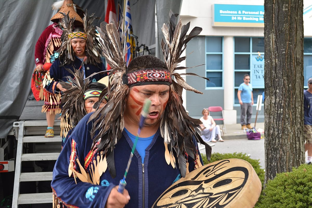 Indigneous dancers descent a stairway and beat hand drums decorated with bird figures, and wearing head dresses full of large feathers.