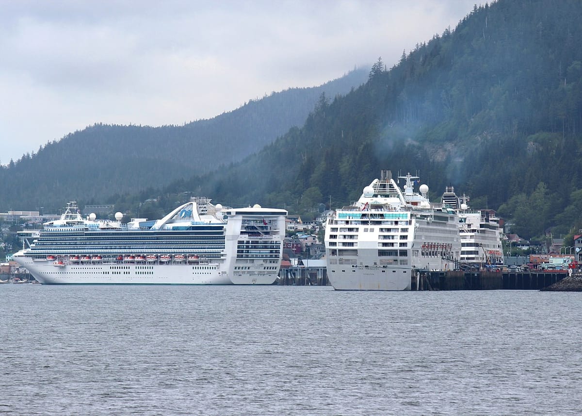 Three massive cruise ships docked at a port surrounded by forested mountains spew pollution from their smoke stacks