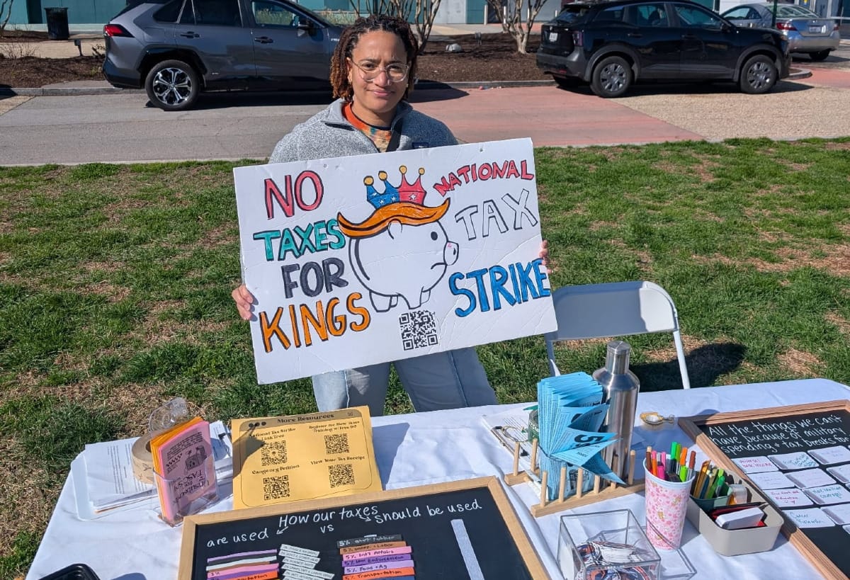 A person at a protest table holds a sign that reads: No Taxes For Kings and National Tax Strike