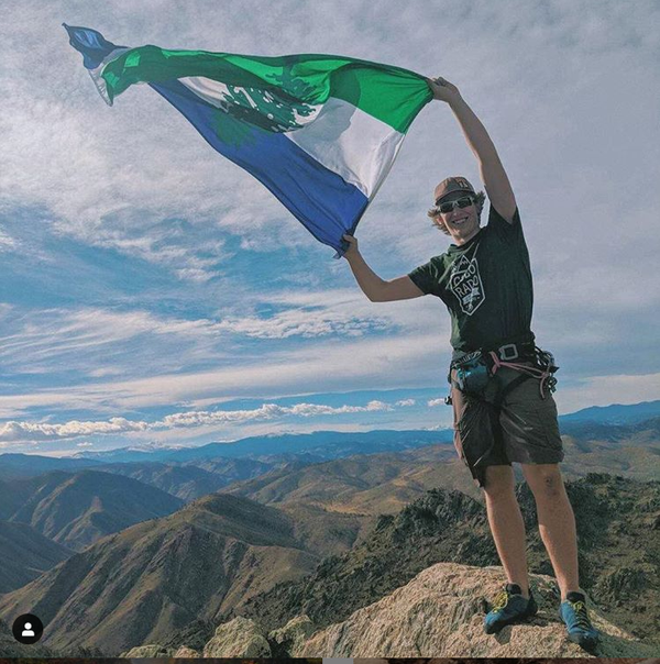 A hiker holds a Cascadia Doug flag in the wind at the top of a peak in the Cacade range.
