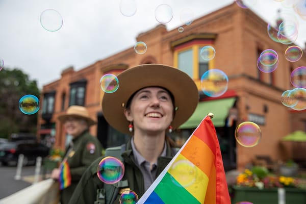 An employee of the National Park service, in uniform, holds a rainbow pride flag and is surrounded by iridescent bubbles.